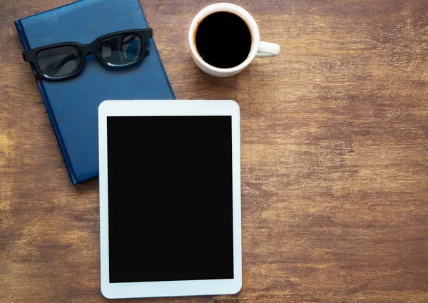 Digital tablet with black blank space on the screen and cup of coffee, Copy space. Closed blue notebook with black glasses on it, top view