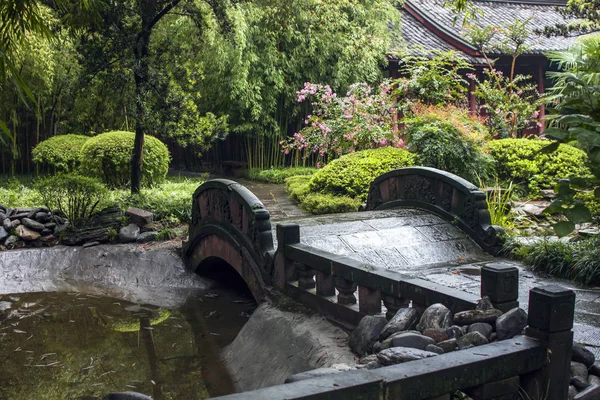 Stone Bridge in a Chinese Garden. Traditional Garden Design with Stone Bridge over a Small Pond Surrounded by Trees and Flowers on a Rainy Day.