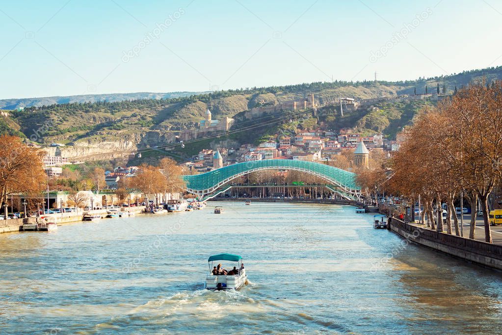 Autumn in Tbilisi, Georgia. Panoramic view over famous glass Bridge of Peace and old town beneath the ancient fortress. Boat riding on the river.