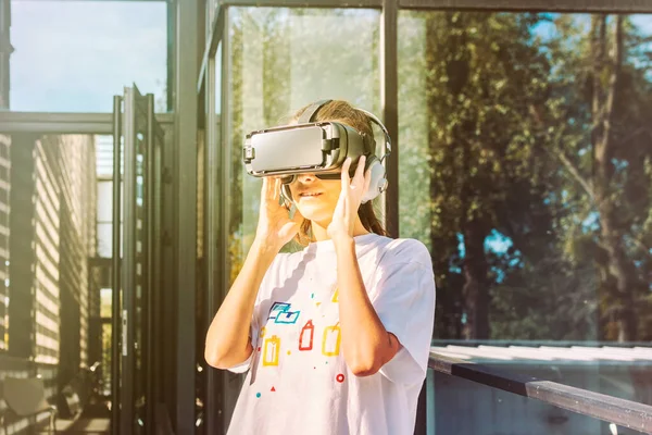 Beyaz tişörtlü güzel bir bayan sanal gerçeklik kulaklığı kullanıyor ellerini yandan tutuyor ve gülümsüyor. Arka planda modern cam iş binası cephesi.