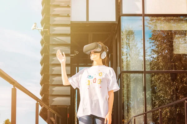 Hermosa joven hembra en camiseta blanca usando auriculares de realidad virtual alcanzando con la mano y sonriendo. Fachada moderna del edificio del negocio del vidrio en el fondo . — Foto de Stock