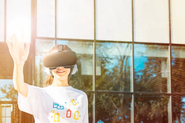 Hermosa joven hembra en camiseta blanca usando auriculares de realidad virtual alcanzando con la mano y sonriendo. Fondo diseñado por VR — Foto de Stock