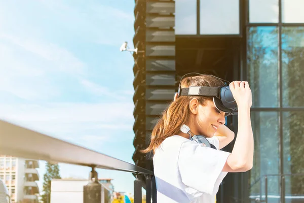 Hermosa joven mujer en camiseta blanca que se pone los auriculares de realidad virtual. Fachada moderna del edificio del negocio del vidrio en el fondo . — Foto de Stock