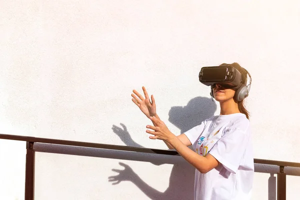 Hermosa joven hembra en camiseta blanca con auriculares de realidad virtual con las manos en el frente. Superficie de pared blanca en el fondo . — Foto de Stock