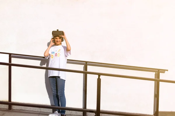 Mulher bonita jovem em t-shirt branca colocando em fone de ouvido realidade virtual e olhando para a câmera com um grande sorriso bonito. Fachada de parede branca no fundo . — Fotografia de Stock