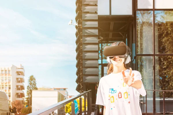 Hermosa joven hembra en camiseta blanca usando auriculares de realidad virtual alcanzando con la mano y sonriendo. Fachada moderna del edificio del negocio del vidrio en el fondo . — Foto de Stock