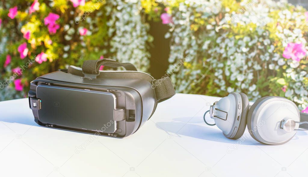 Virtual reality headset equipment with high quality wireless blootooth headphones for complete virtual experience. White table surface with garden flowers in the background. Bright sunny daytime.