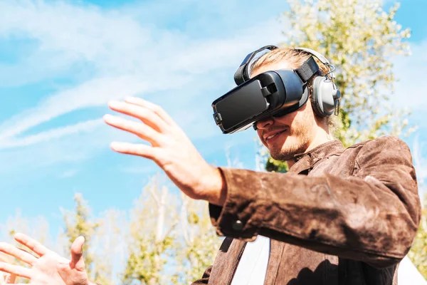 Uzun saçlı, sanal gerçeklik gözlüğü takan ve dışarıda kulaklık takan genç bir erkek. Vr gözlüklü akıllı telefon..