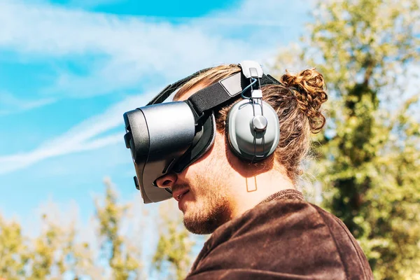 Joven hombre con el pelo largo con gafas de realidad virtual y auriculares grandes al aire libre. Smartphone con gafas VR auriculares . — Foto de Stock