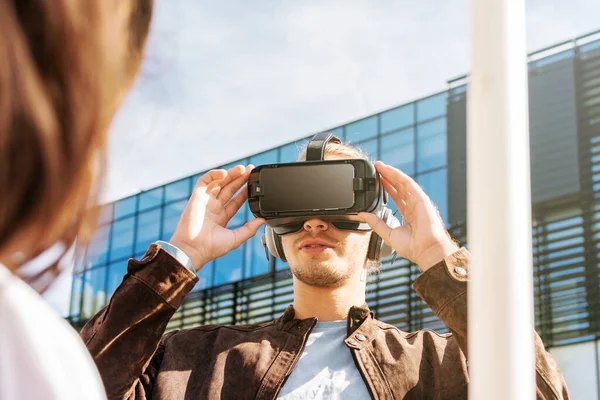 Joven hombre con el pelo largo con gafas de realidad virtual y auriculares grandes al aire libre. Smartphone con gafas VR auriculares. Sostener las gafas con las manos levantadas. Edificio de negocio de vidrio en el backgrou — Foto de Stock