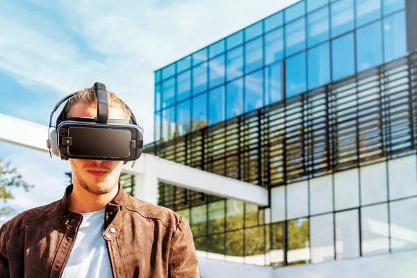 Joven hombre con el pelo largo con gafas de realidad virtual y auriculares grandes al aire libre. Smartphone con gafas VR auriculares. Edificio de negocio de vidrio en el fondo . — Foto de Stock