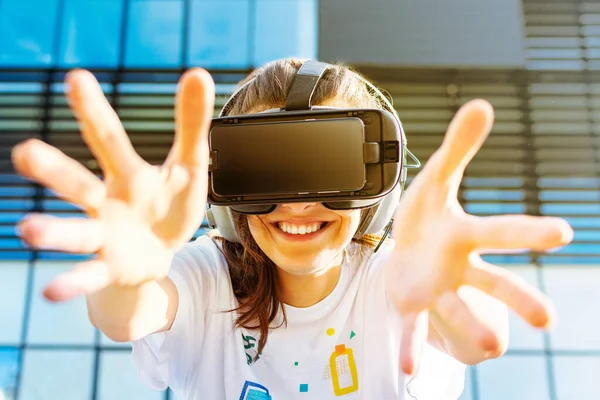 Mujer joven con el pelo largo con gafas de realidad virtual y auriculares grandes al aire libre. Amplia sonrisa y las manos abiertas hacia la pantalla. Smartphone con gafas VR auriculares. Edificio de negocio de vidrio en la parte trasera — Foto de Stock