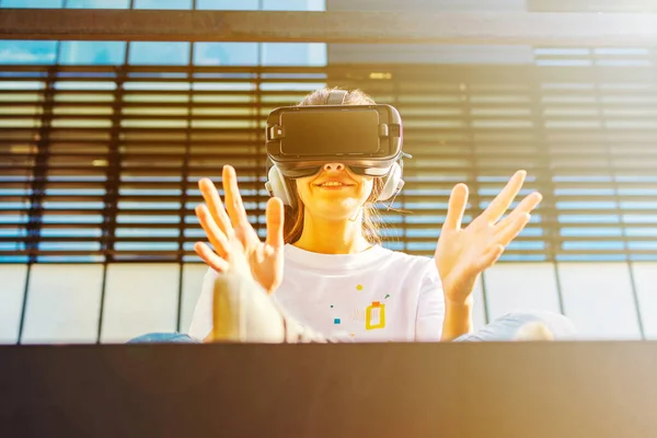 Mujer joven con el pelo largo sentado en el suelo y con gafas de realidad virtual y auriculares grandes al aire libre. Amplia sonrisa y las manos abiertas hacia la pantalla. Smartphone con gafas VR auriculares. Bus de vidrio — Foto de Stock