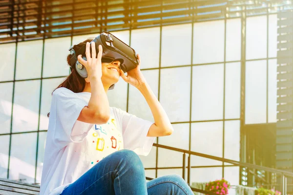 Mujer joven con el pelo largo sentado en las escaleras y con gafas de realidad virtual con auriculares grandes. Amplia sonrisa y las manos sosteniendo el dispositivo. Smartphone con gafas VR auriculares. Negocio del vidrio b — Foto de Stock