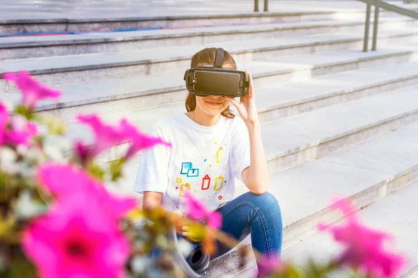 Mujer joven con el pelo largo sentado en las escaleras y con gafas de realidad virtual con auriculares grandes. Amplia sonrisa y las manos sosteniendo el dispositivo. Smartphone con gafas VR auriculares. Negocio del vidrio b — Foto de Stock