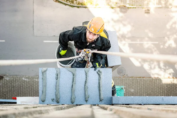 Male worker hanging on rope access and height construction project. Working on building wall insulation and facade painting decoration job. Looking up to camera