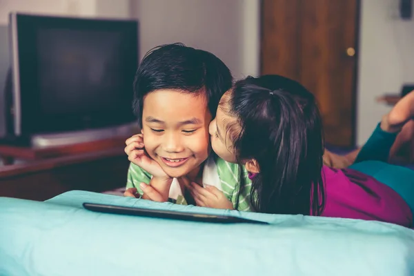 快乐的亚洲儿童微笑着使用数码片 姐姐亲吻她的弟弟的脸颊 躺在床上容易 爱的兄弟姐妹的概念 老式胶片过滤效果色调 电影风格 — 图库照片