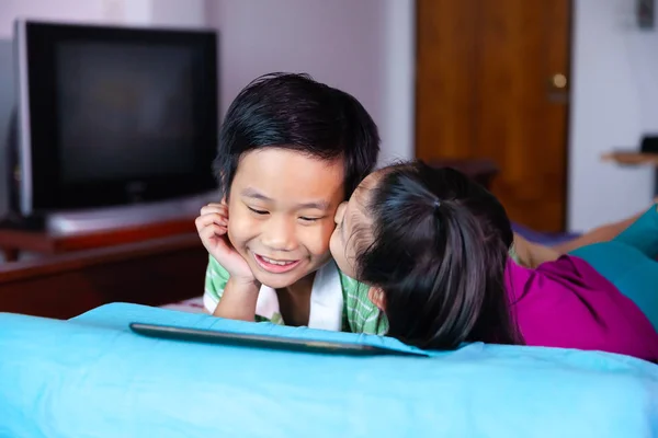 快乐的亚洲儿童微笑着使用数码片 姐姐亲吻她的弟弟的脸颊 躺在床上容易 关爱兄弟姐妹的概念 — 图库照片
