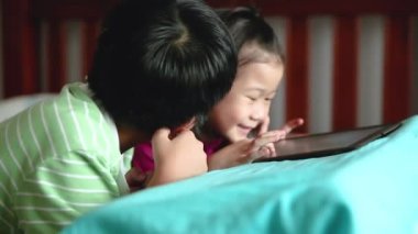Asya çocuk dijital tablet kullanma. Gülümseyen ve yakınındaki kız kardeşi tarafından tezahürat kardeşi mutlu. Heyecanla touchpad üzerinde oyun ve yatakta yüzükoyun yatarken şirin kız. Çocuktan kıza değişen odak.