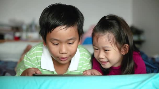 アジアの子供たちはデジタル タブレットを使用します 笑顔と近くで兄を応援して喜んで姉妹 かわいい少年はタッチパッドで興奮してゲームをプレイしやすいベッドに横たわっています — ストック動画