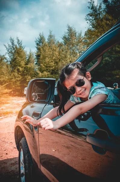 Cute asian girl in sunglasses smiling with perfect smile while sitting in the car. Tourist child relaxing, outdoors at the daytime with bright sunlight on summer day. Travel on vacation concept.