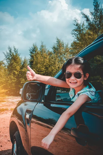 Cute asian girl in sunglasses smiling with perfect smile while sitting in the car. Tourist child relaxing and showing thump up. Outdoors with bright sunlight on summer day. Travel on vacation concept.