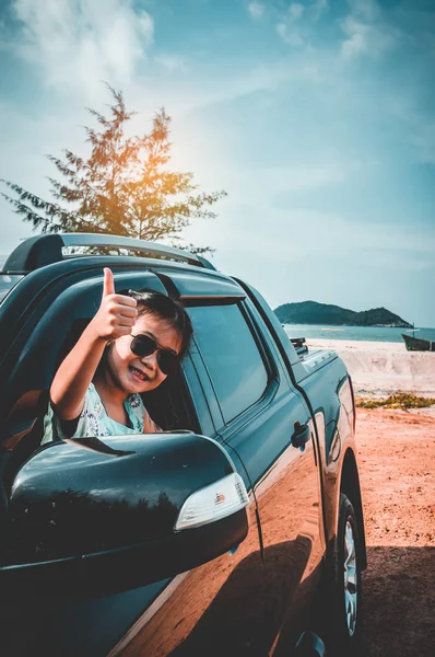 Cute asian girl in sunglasses smiling with perfect smile at beach while sitting in the car. Tourist child relaxing and showing thump up. Outdoors with bright sunlight on summer day. Travel on vacation