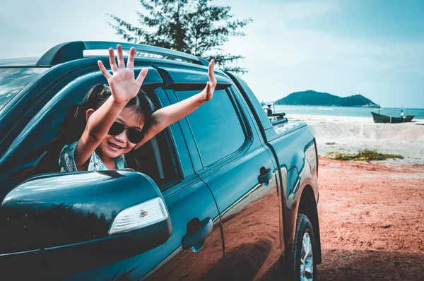 Cute asian girl in sunglasses smiling with perfect smile at beach while sitting in the car. Happy child raised her arms up for enjoy. Outdoors with sunlight on summer day. Travel on vacation concept.