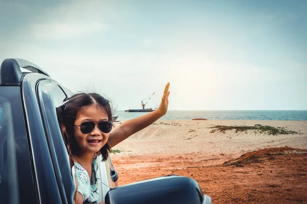 Happy asian girl in sunglasses smiling with perfect smile at beach while sitting in car. Cute child raised arms up for enjoy. Outdoor on summer day. Travel on vacation concept. Teal and orange filter.
