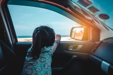 Arkadan görünüm. Mutlu Asyalı kız arabada otururken kumsalda zevk. Çocuk gözlem doğadan penceresini açın. Yaz gününde güneş ışığı ile açık. Tatil kavramı üzerinde seyahat. Yeşil ve turuncu filtre