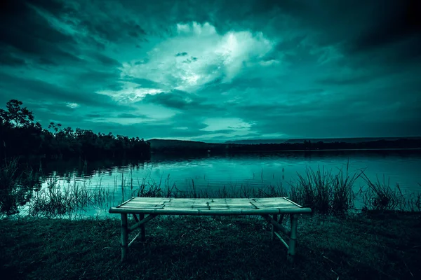 Sampah bambu di rumput di depan danau dan cahaya bulan di belakang awan di malam hari di atas danau yang tenang . — Stok Foto