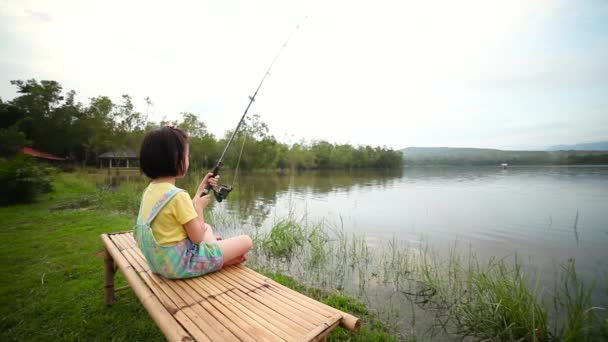 竹のごみの上に座って素敵なアジアの子供 湖の風景に対して釣りながら 幸せな女の子は彼女の手の中にロッドを引く 自然と子どもをつなぐというコンセプト 休暇中の旅行 — ストック動画