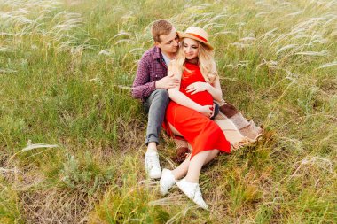 Hamile bir kız, karnından doğumu bekliyor, kırmızı elbiseli ve hasır şapkalı, kocasıyla birlikte tarlada ve tüy yumağı ile.