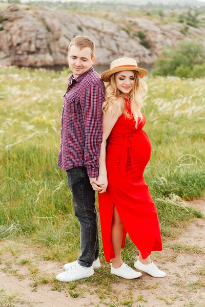 赤いドレスと草原と羽の草の彼女の夫とのわらの帽子で 出産を待っているおなかを持つ妊婦 — ストック写真