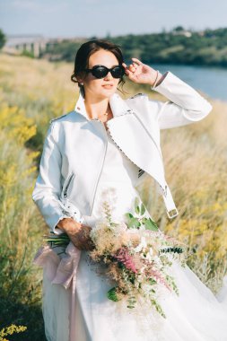 Şık, beyaz bir gelinlik içinde, deri ceketli ve güneş batarken gözlüklü gelin.