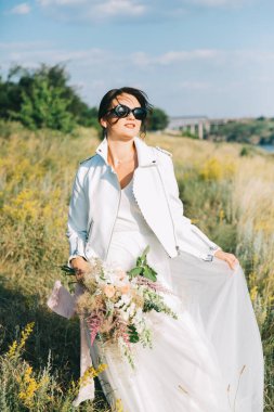 Şık, beyaz bir gelinlik içinde, deri ceketli ve güneş batarken gözlüklü gelin.