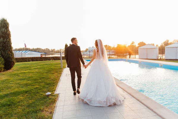 Güzel, lüks bir çift, siyah takım elbiseli damat ve duvağı ve beyaz gelinliği olan gelin, el ele tutuşmuş, rıhtımda havuz kenarında yürüyorlar, yat ve geminin yanında.