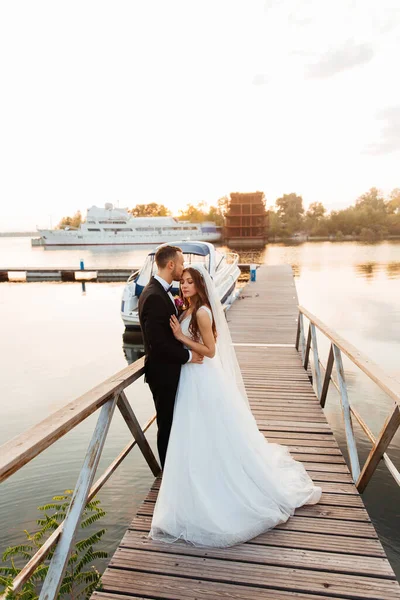 美しい豪華なカップル 黒いスーツの新郎とベールと白いウェディングドレスの花嫁 手を握り 桟橋のプールで歩く ヨットと船の隣 — ストック写真