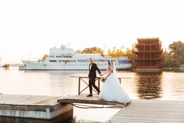 美丽的一对豪华夫妇 穿着黑色西服的新郎 戴着面纱和白色婚纱的新娘 手牵手 走在码头上的游泳池边 坐在游艇和船旁边 — 图库照片