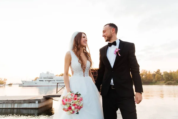 美しい豪華なカップル 黒いスーツの新郎とベールと白いウェディングドレスの花嫁 手を握り 桟橋のプールで歩く ヨットと船の隣 — ストック写真
