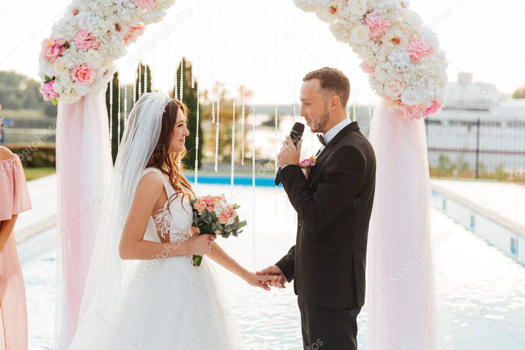 Luxurious beautiful couple, the groom in a black suit and the bride in a wedding white dress, exchanged rings at an exit ceremony with an arch, wedding in the fall