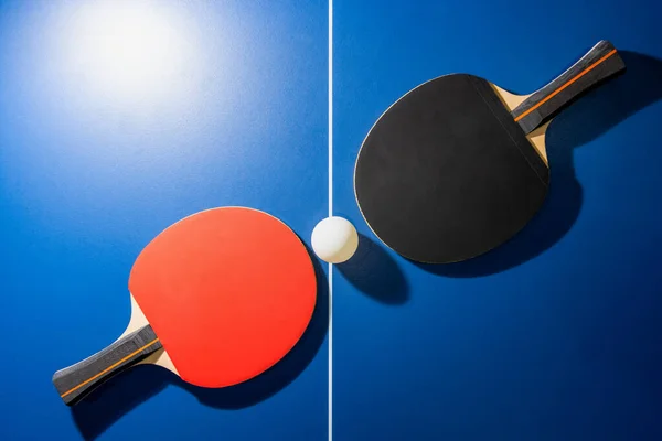 Üst manzara siyah ve kırmızı masa tenisi raketi ve mavi masa tenisi masasında parlak bir spot ışığı olan beyaz ping pong topu, iki masa tenisi raketi kapalı alanda egzersiz yapmak için bir spor yarışması ekipmanıdır.
