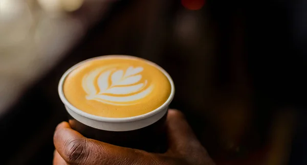 African Coffee Barista holding a take away cup with milk foam in a leaf shape