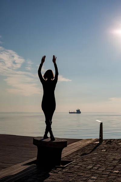 在希腊塞萨洛尼基的停靠杆上 跳舞的年轻漂亮合身的女子的背部观看 后视图与蓝色海和天空和船在背景 — 图库照片