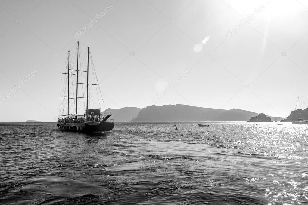 Tourist boat sailing away in Santorini, Greece. Amazing daytime view towards the Sun and deep sea crystal waters, bright black and white image with a lot of light