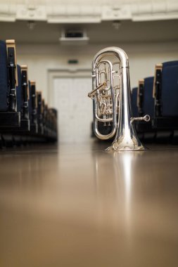 A euphonium standing on its bell on the floor amongst rows of chairs clipart