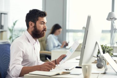 Sakallı adam modern ışık Office belgeleriyle çalışırken bilgisayar izlerken yan görünüm