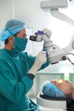 Asyalı Doktor mikroskop işletim sistemi çalıştıran olgun hastanın gözünde keşfetmek bir cerrah her bir üniformalı erkeklere