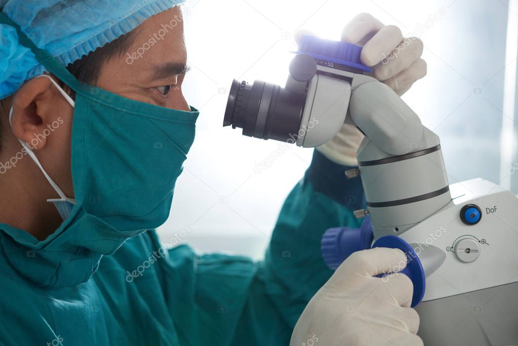 Asian doctor in mask looking at lens of operating microscope during surgery