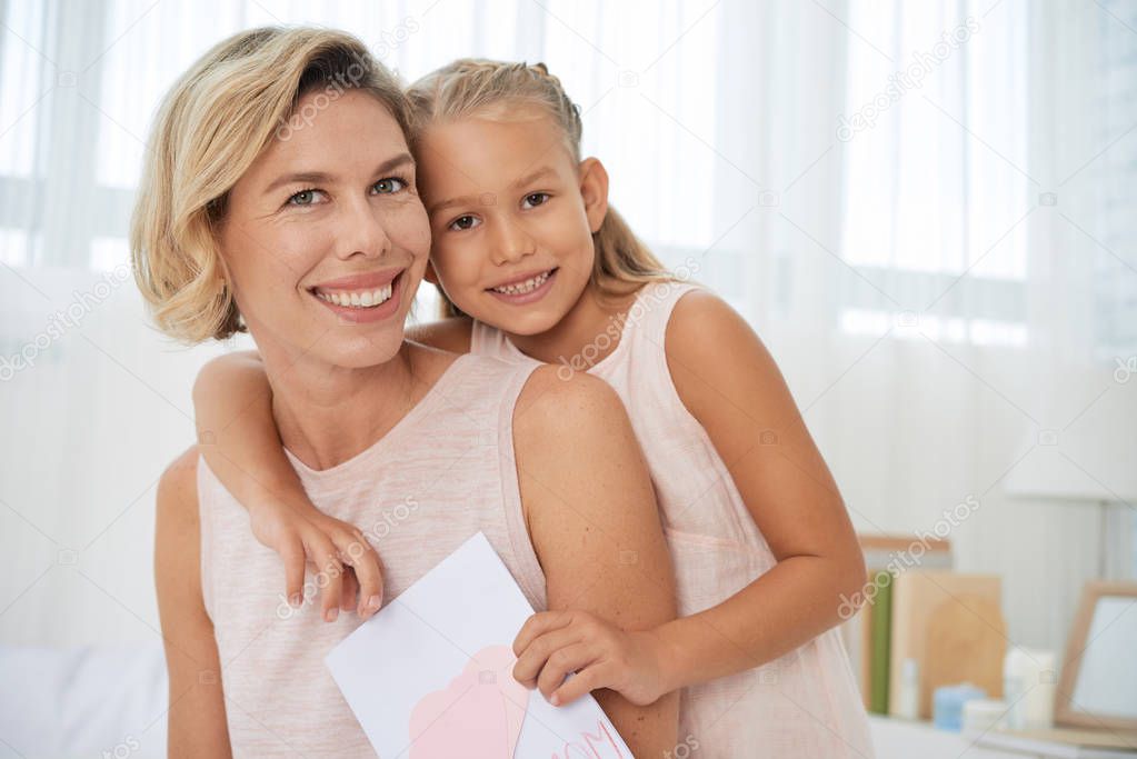 Portrait of happy woman and adorable little daughter holding greeting card and looking at camera 